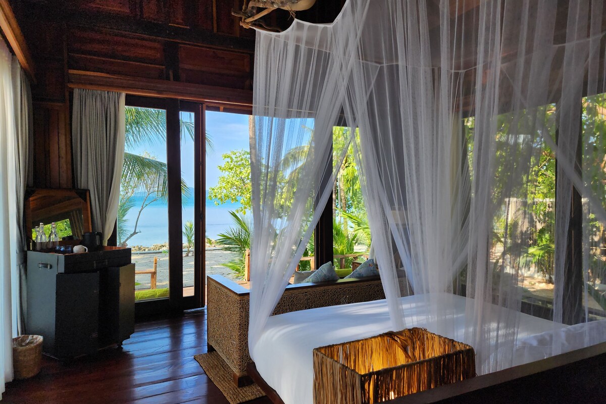A bedroom with a wooden floor, a bed with a canopy, a bedside table, and a view of the beach and ocean.