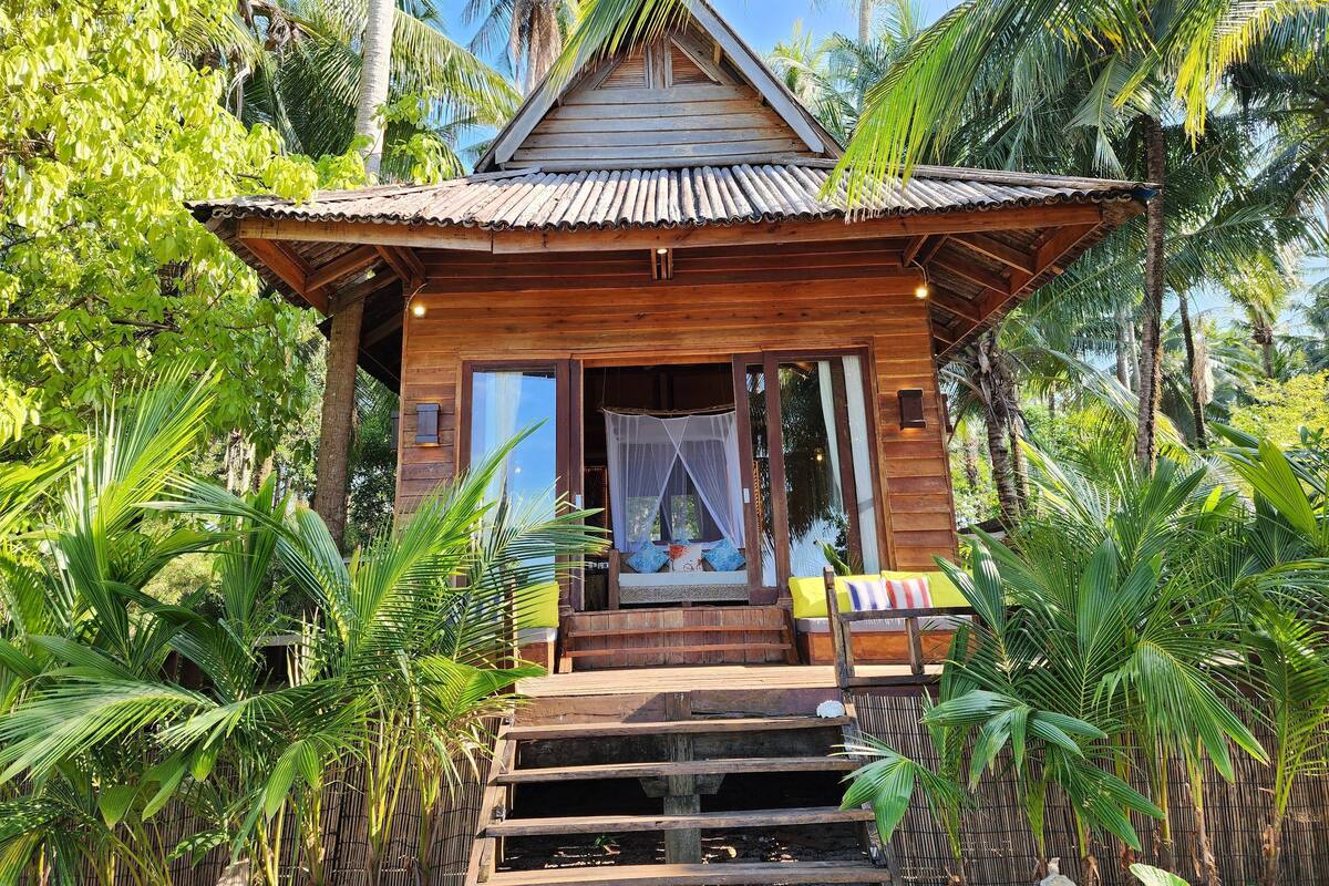 A wooden bungalow with a thatched roof, surrounded by lush tropical vegetation.
