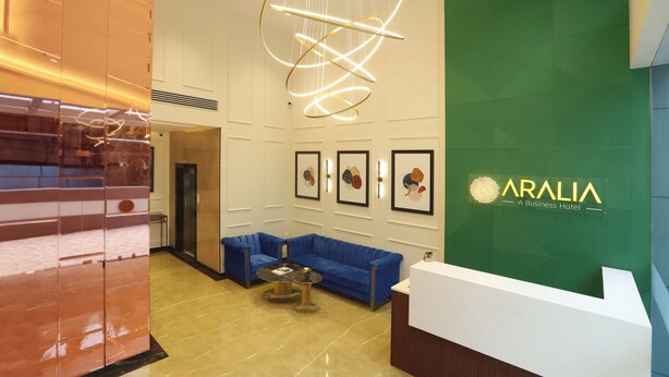 A reception area with a blue sofa, a round table, and a green wall with the hotel name. - Aralia - A Business Hotel MIDC (Mumbai)