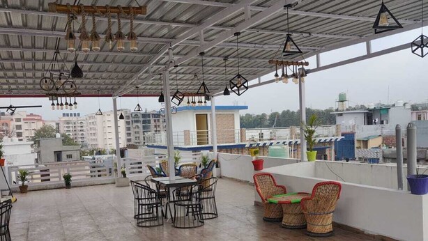 Een terras met tafels en stoelen, een hoog plafond met hangende lampen, en een uitzicht op gebouwen op de achtergrond.