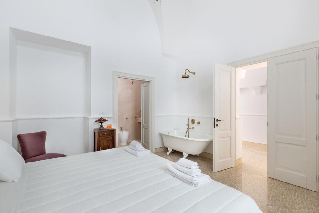 A hotel room with a large bed, a red chair, a small table with a lamp, a white bathtub, and a white door leading to another room.