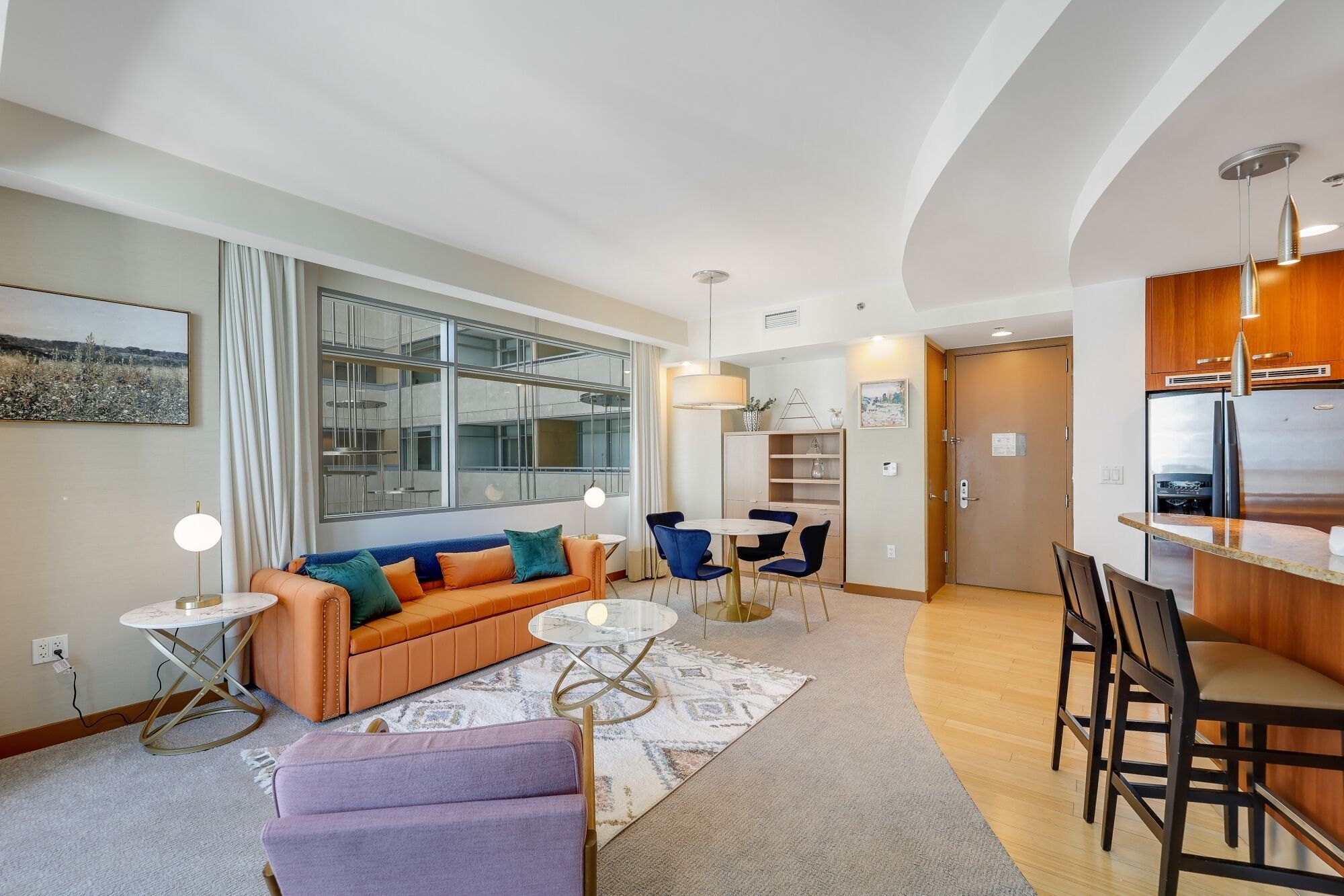 A modern living room with a sofa, coffee table, and chairs, a dining area with a table and chairs, and a kitchen with a refrigerator and wooden cabinets.
