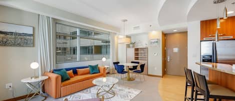 A modern living room with a sofa, coffee table, and chairs, a dining area with a table and chairs, and a kitchen with a refrigerator and wooden cabinets.