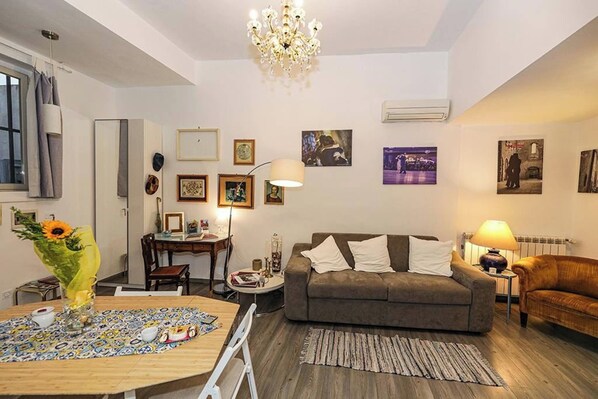 A living room with a sofa, a dining table with a vase of flowers, a chandelier, and framed pictures on the wall.