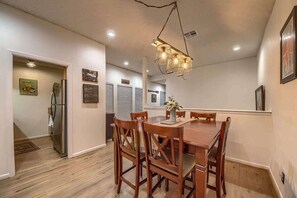 A dining area with a wooden table and chairs, a hanging light fixture, and a refrigerator in the background.