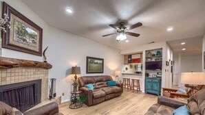 A living room with a fireplace, leather sofas, a ceiling fan, and a kitchen area in the background. - CW B201 Comal Riverbahn Retreat B201 (New Braunfels)