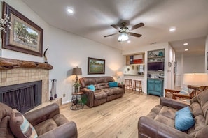 A living room with a fireplace, leather sofas, a ceiling fan, and a kitchen area in the background.