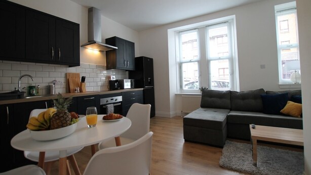 Een moderne keuken met donkere kasten, een wit aanrechtblad en een eetgedeelte met een tafel en stoelen. Er is een grijze bank en een raam met zonwering in de woonkamer.