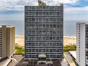 Un hotel a più piani con una moderna facciata in vetro, situato vicino a una spiaggia con parcheggio ben visibile e una vista aperta sull'oceano.