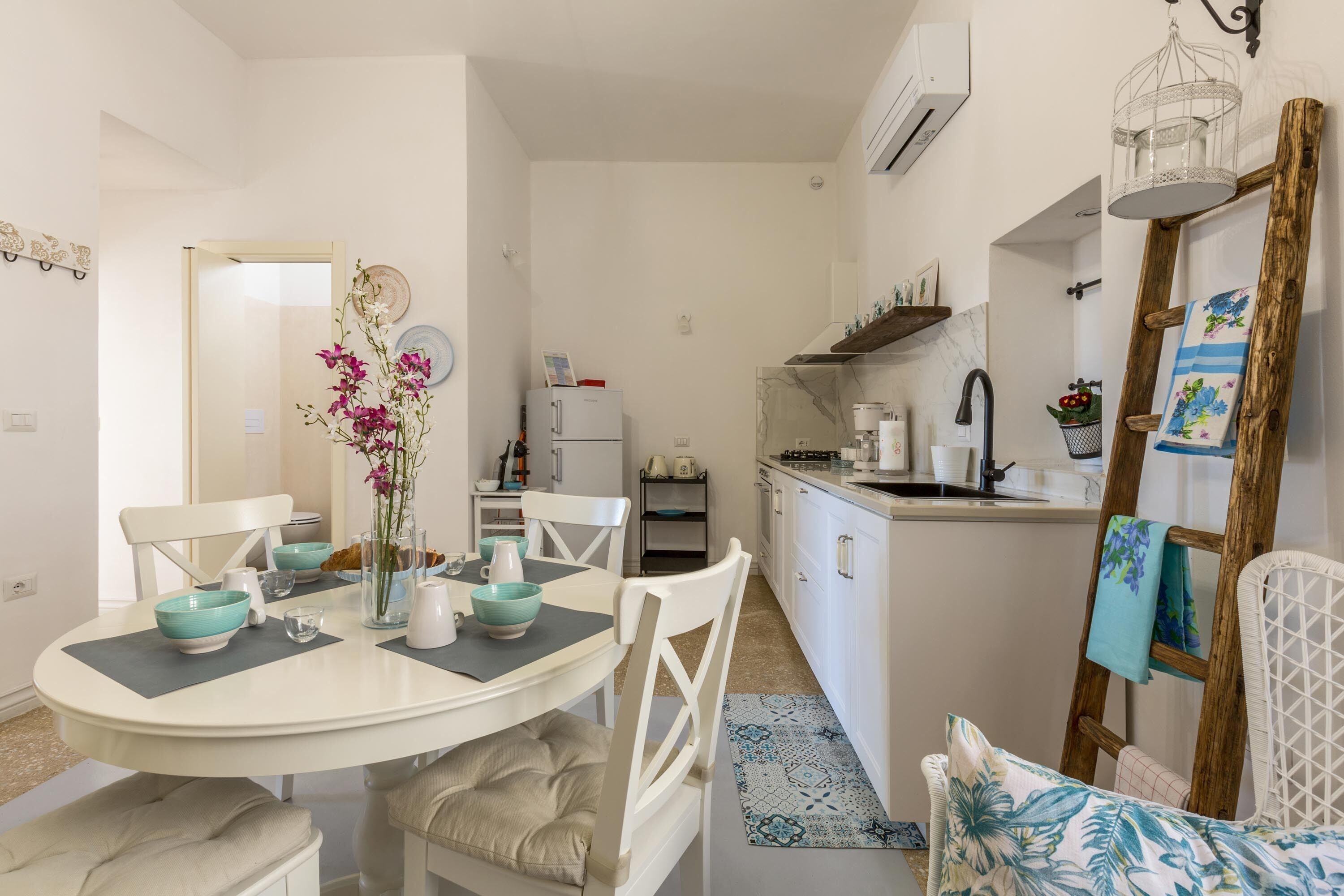 A compact kitchen with white cabinets, a white refrigerator, and a sink. There is a dining table with teal bowls and a vase of flowers.