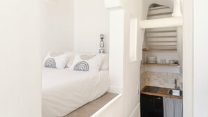 A bedroom with a bed, pillows, a nightstand, a wall-mounted lamp, and a kitchen area with a sink and shelves. - Le Mini - Un Voyage en Provence (Aix-en-Provence)