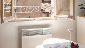 A room with a large open window, a radiator, a round table with a book and a bowl of fruit, and a white wall. - Le Mini - Un Voyage en Provence (Aix-en-Provence)