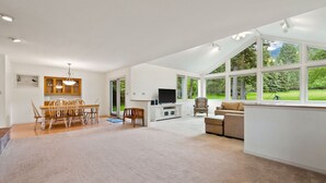 A spacious living room with a large window, a dining area with a wooden table and chairs, and a television on a white stand.