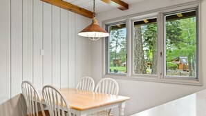 A dining area with a wooden table and white chairs, a hanging pendant light, and a window with a view of greenery.