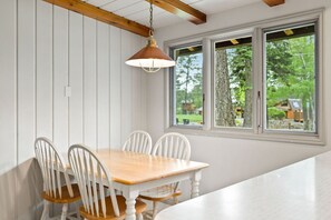 A dining area with a wooden table and white chairs, a hanging pendant light, and a window with a view of greenery.