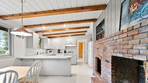 A modern kitchen with a central island, wooden beams, a brick fireplace, and a dining area with white chairs.