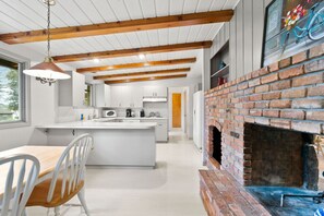A modern kitchen with a central island, wooden beams, a brick fireplace, and a dining area with white chairs.