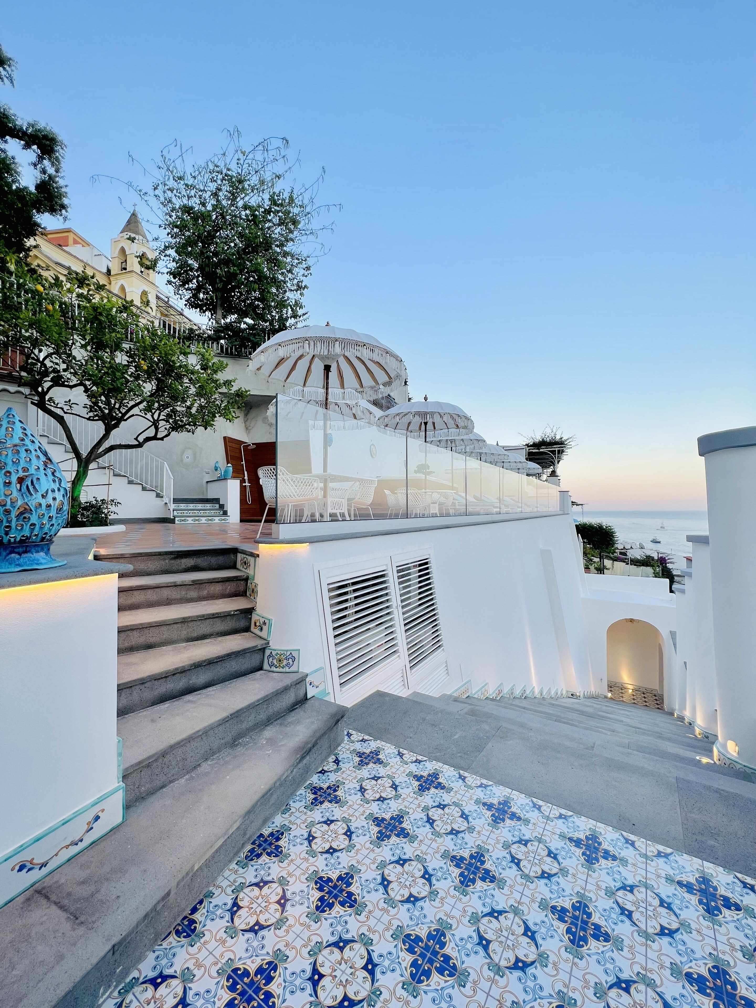 A staircase leading to a white building with a dome structure, a tree, and a view of the sea.