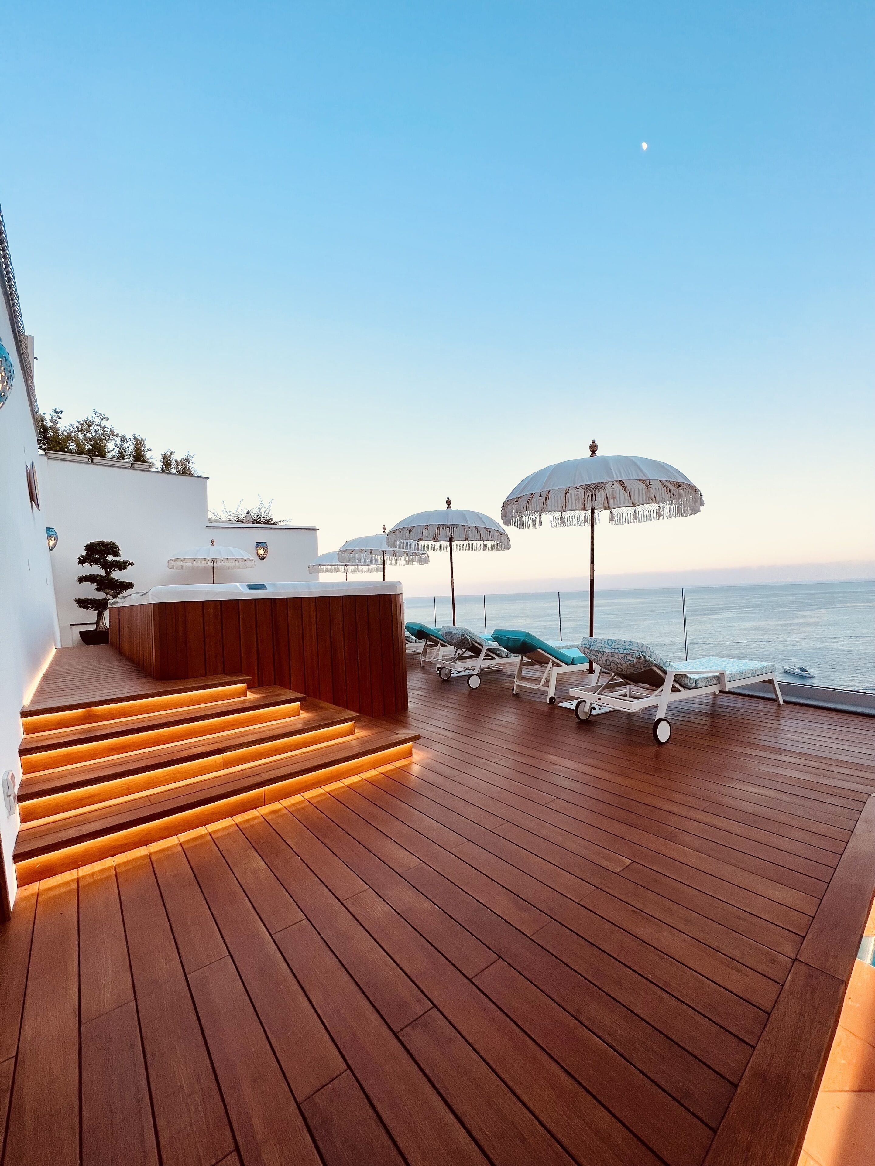 A wooden deck with a jacuzzi, lounge chairs, and umbrellas overlooking the sea.