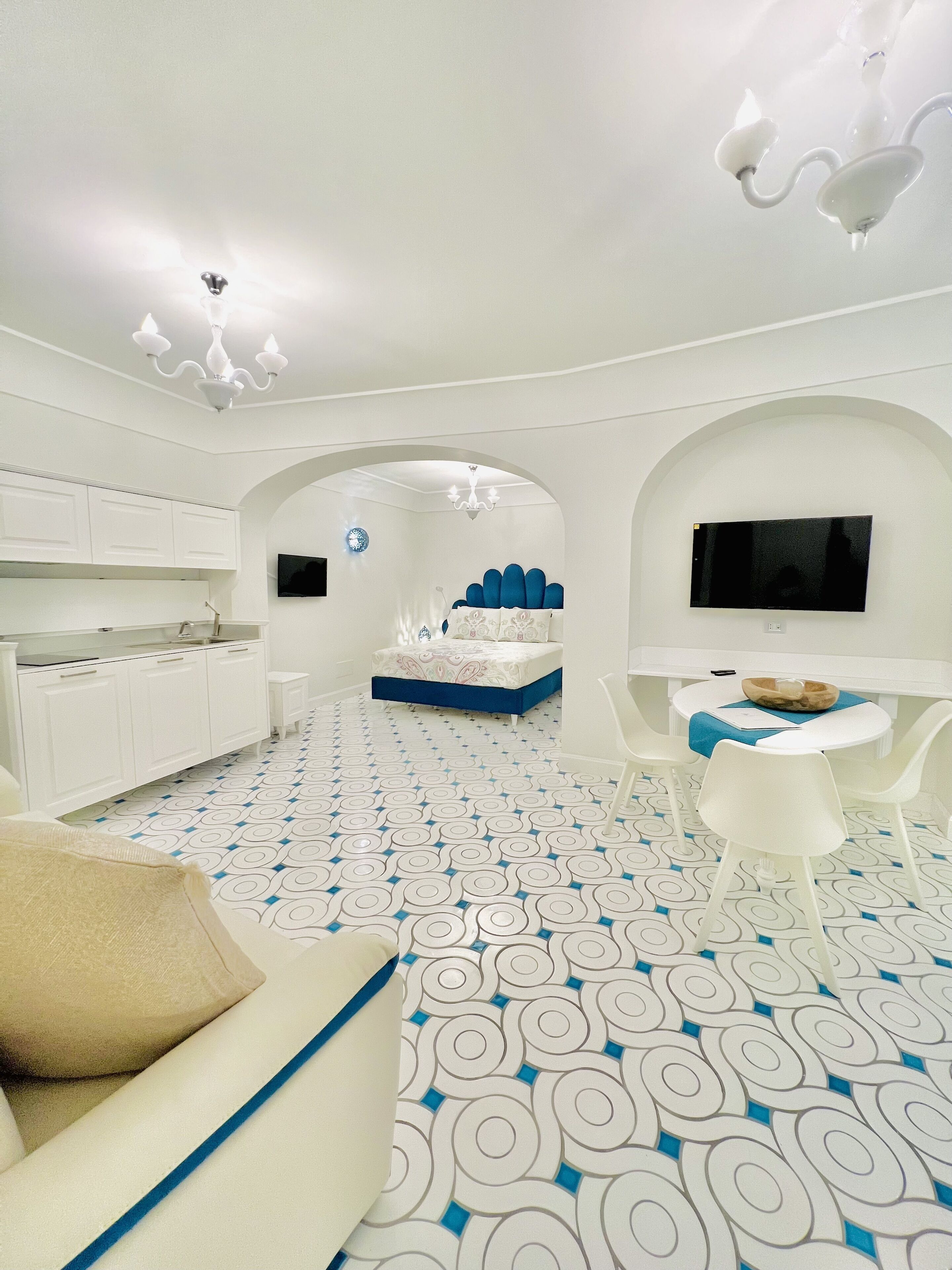 Ein modernes, weißes Zimmer mit gemustertem Boden, einer Couch, einem Essbereich mit Stühlen, einem Bett mit blauer Kopfteil und einem an der Wand montierten Fernseher.
