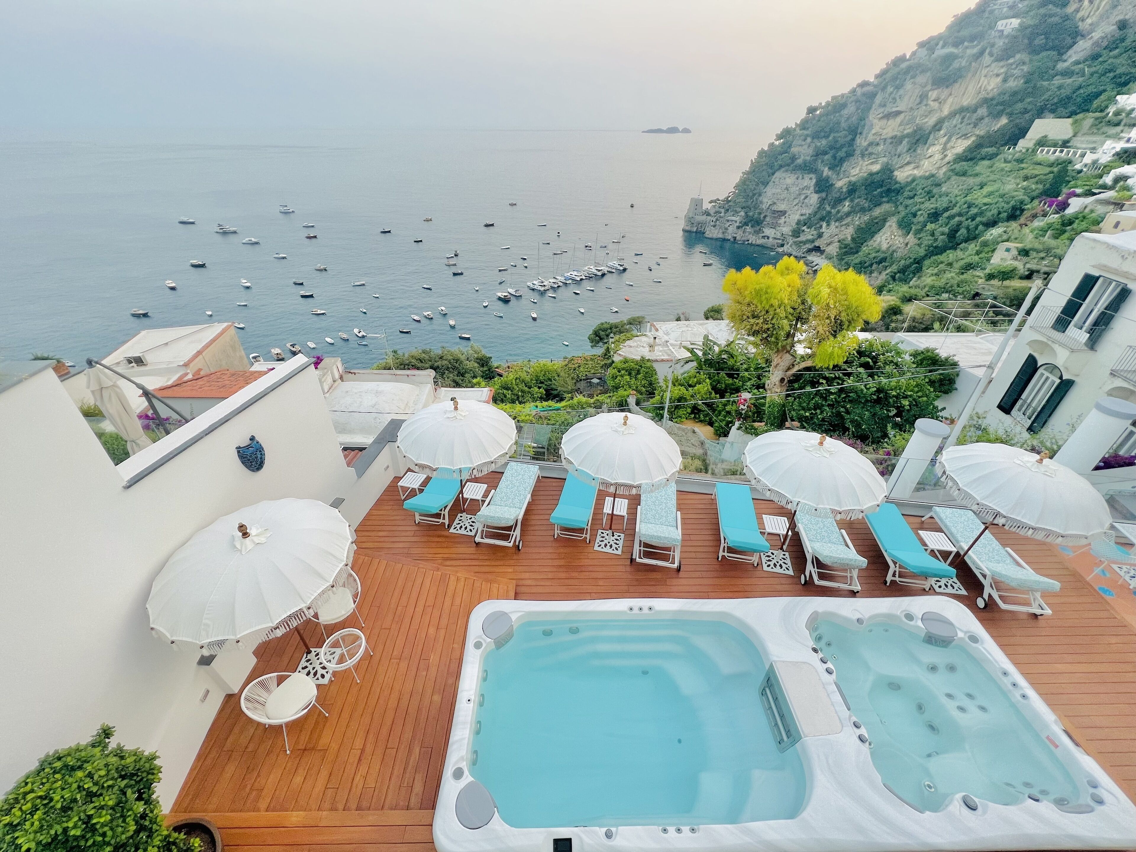 A rooftop terrace with a hot tub, lounge chairs, and umbrellas overlooking a coastal area with boats.