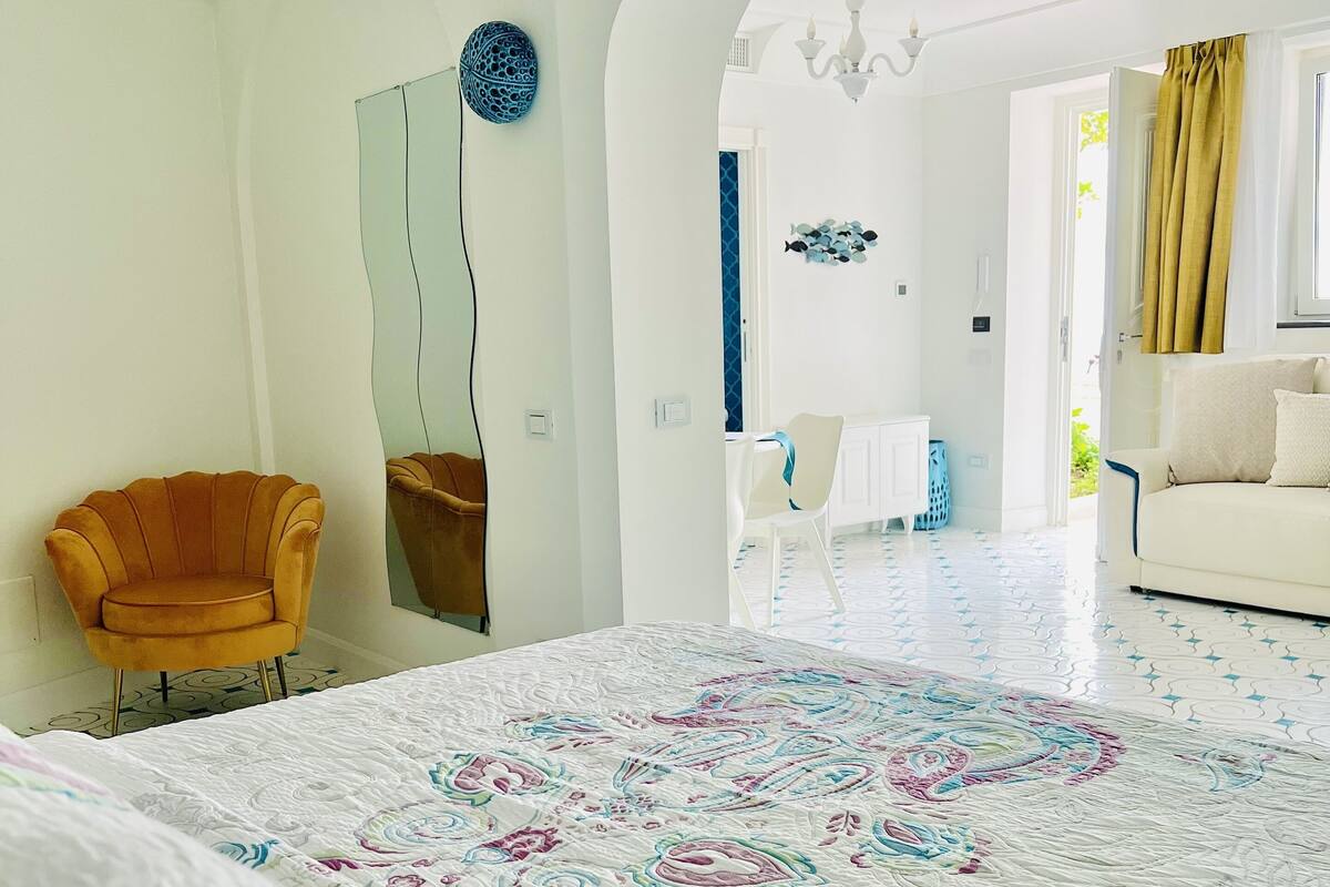Ein Schlafzimmer mit einem großen Bett, einem Sessel, einem Spiegel und Blick in einen anderen Raum.