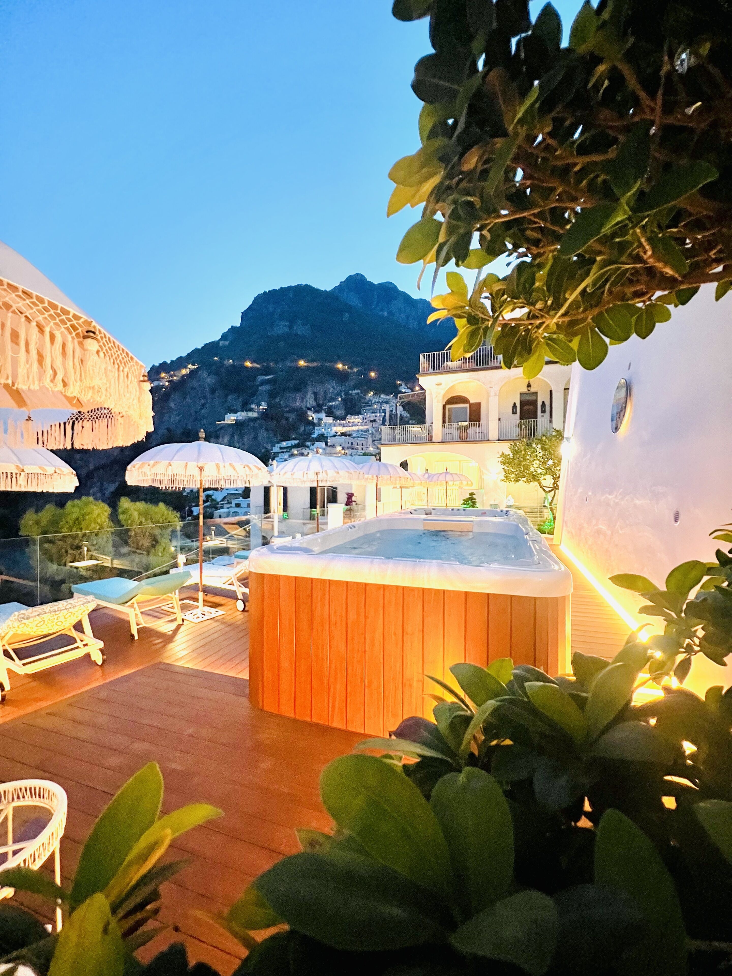 A hot tub on a wooden deck with lounge chairs, umbrellas, and a mountain backdrop.