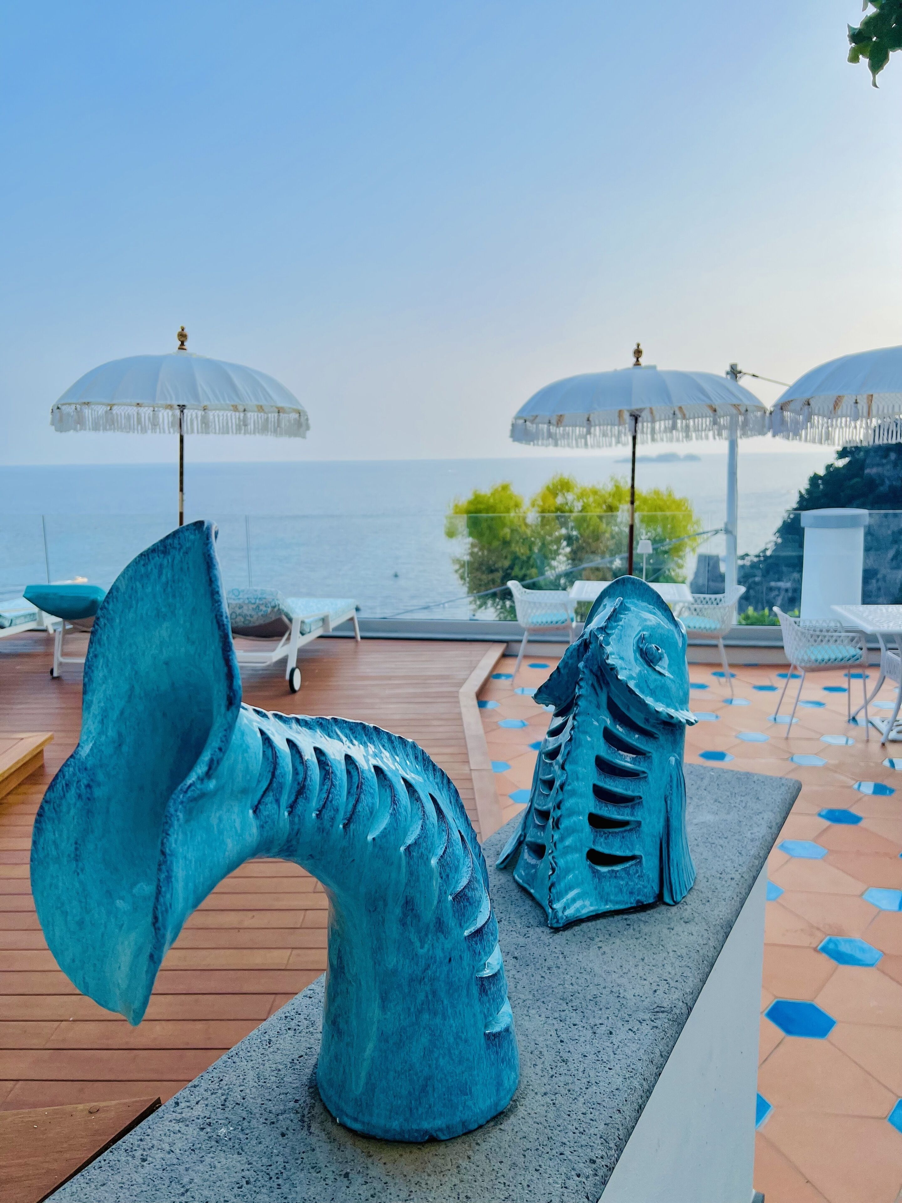 Una terraza con dos esculturas de peces azules, sombrillas blancas y vista al mar.