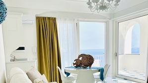 A modern living room with a white sofa, a dining table with chairs, a chandelier, and a view of the sea through large windows.