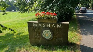 Ein Schild mit dem Namen 'MASHAM' und einem Wappen, umgeben von Grünpflanzen und einer Straße.