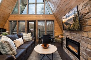 A cozy interior with a fireplace, a wooden ceiling, and a large window overlooking a forest.