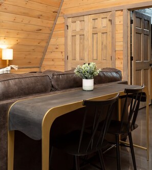 Una habitación acogedora con techo de madera, un sofá de cuero marrón, una pequeña mesa de comedor con dos sillas y una planta en maceta.