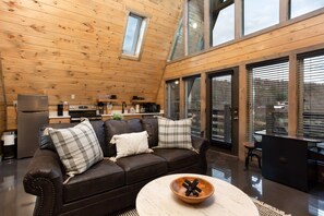 A cozy cabin interior with a wooden ceiling, a sofa with cushions, a coffee table, and a kitchen area with appliances.