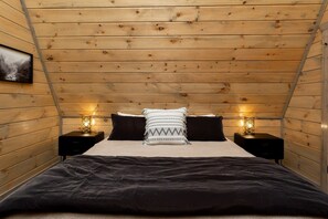 A bedroom with a wooden ceiling, a bed with black bedding, two bedside tables with lamps, and a wall-mounted picture.