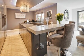 A modern kitchen with a central island, a dining area with a bar stool, and a large window providing natural light. - Host & Stay | Casa Feliz (Skirlaugh)