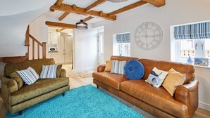 Un salon avec un canapé en cuir marron, un canapé vert et un tapis bleu. Le plafond est en bois et il y a une horloge au mur.