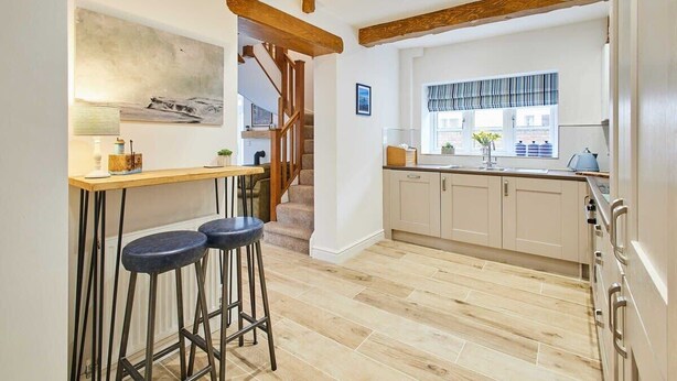 Une cuisine avec un plancher en bois, un coin bar avec des tabourets et une fenêtre agrémentée d’une lambrequin à rayures.