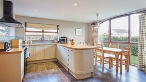 A modern kitchen with a large island, a dining area with wooden chairs, and a view of the outdoors through large windows. - Host & Stay | The Coach House (Beal)