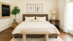 A bedroom with a large bed, a potted plant, two framed artworks, a bedside table with a lamp, and a window with curtains.