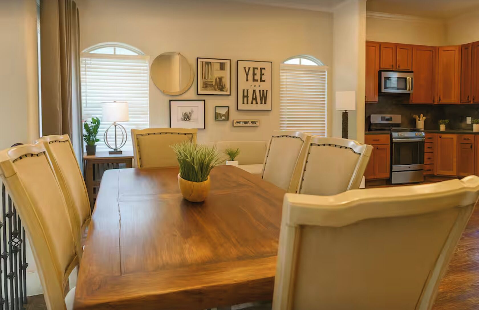 A dining room with a wooden table, beige chairs, a potted plant, framed artwork, and a kitchen in the background.