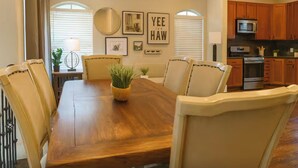 A dining room with a wooden table, beige chairs, a potted plant, framed artwork, and a kitchen in the background.
