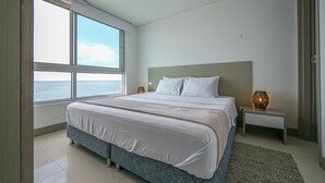 A bedroom with a large bed, a nightstand, a lamp, and a view of the sea through large windows. - Palmetto Beach 1402 & Sea View - Hot Water- 2bth (Cartagena)