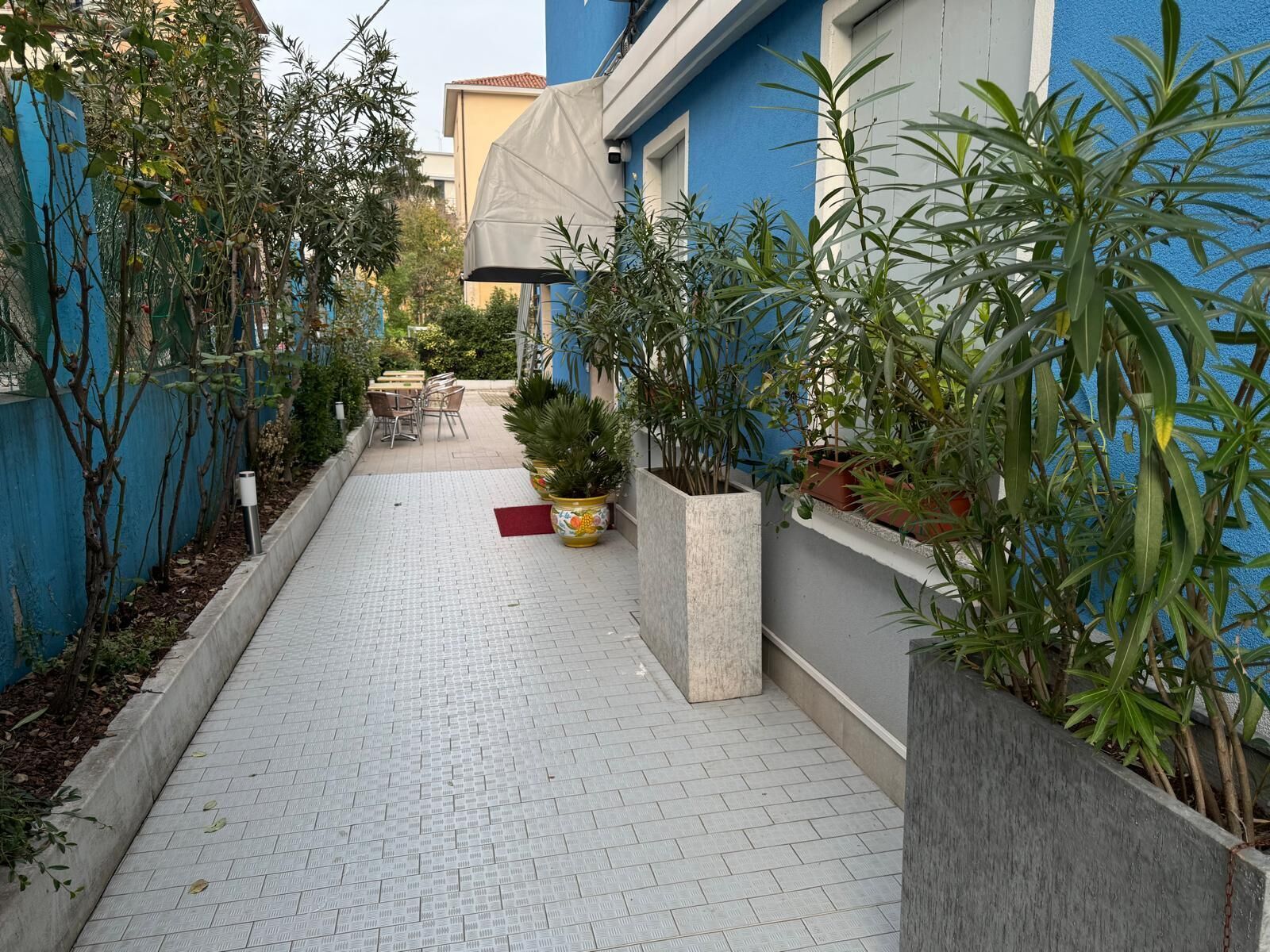 A narrow pathway with tiled flooring, flanked by potted plants and a seating area with chairs and a table.