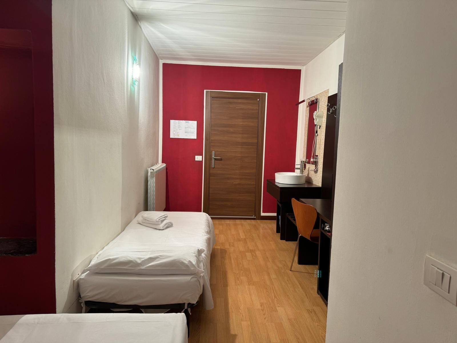 A narrow hotel room with a single bed, a wooden door, a red wall, a small bathroom with a sink and mirror, and a radiator.