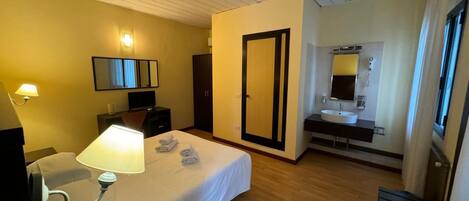 A hotel room with a bed, bedside lamp, a desk with a chair, a wardrobe, a bathroom with a sink and mirror, and a door leading to another room.