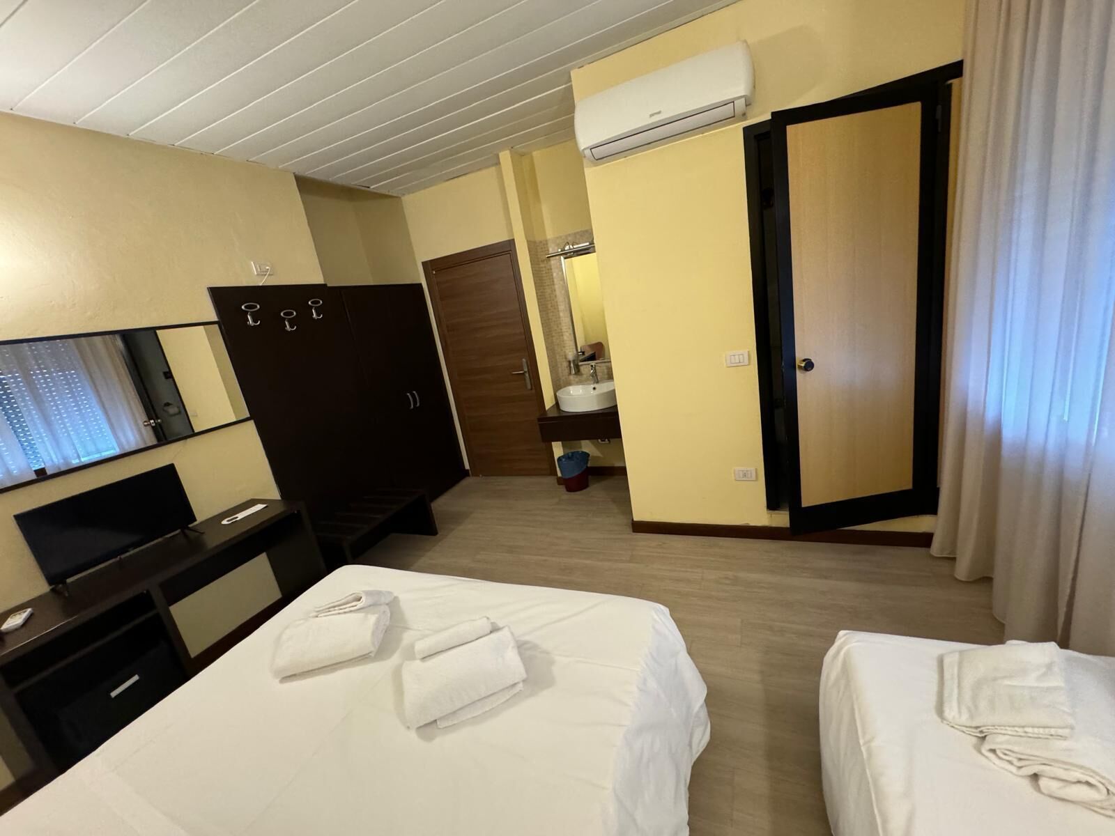 A hotel room with two beds, a desk with a TV, a wardrobe, a bathroom with a sink and toilet, and an air conditioning unit.