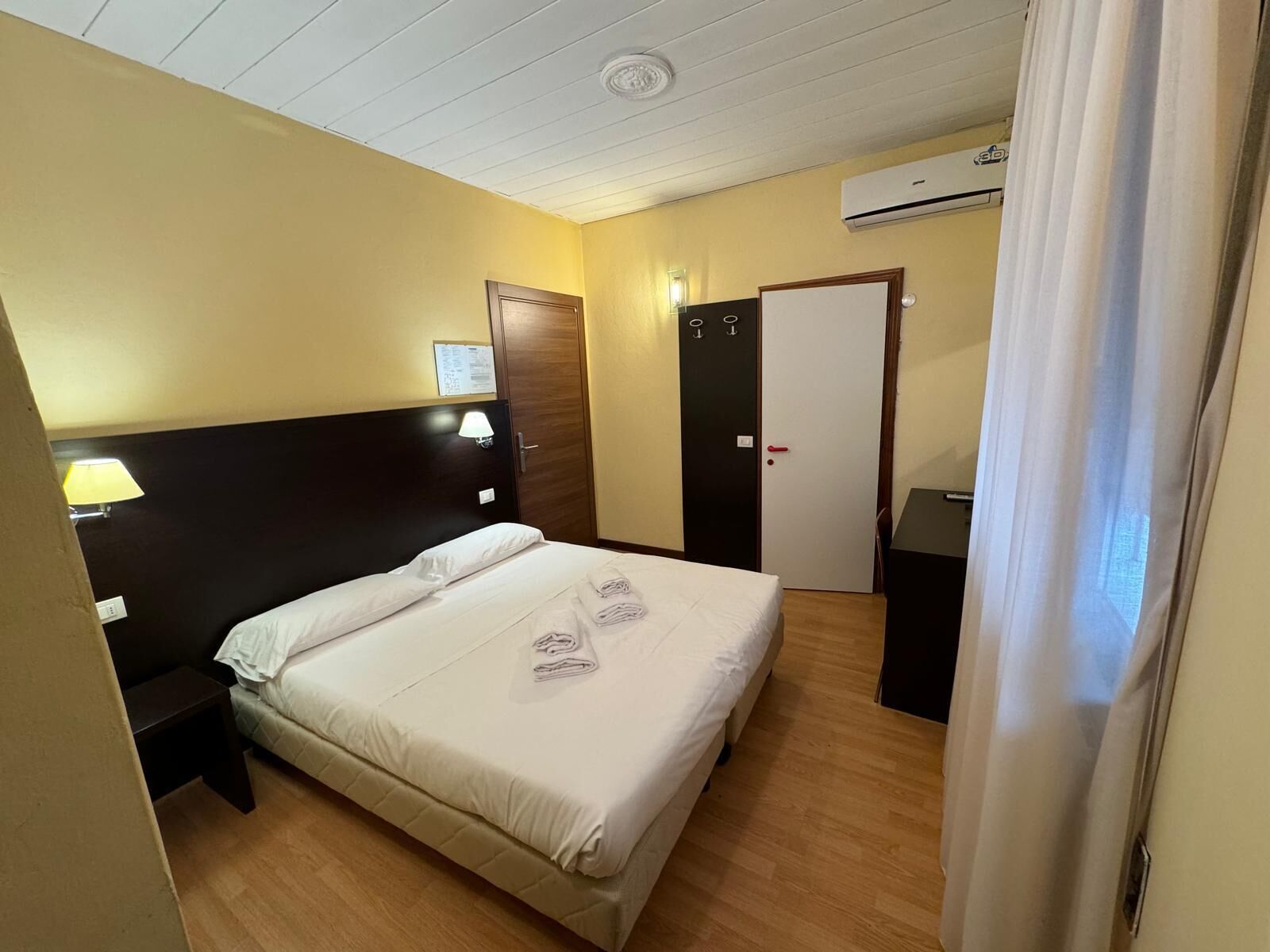 Ett hotellrum med en säng, ett nattduksbord, ett skrivbord, en stol, en dörr och en luftkonditioneringsanläggning.
