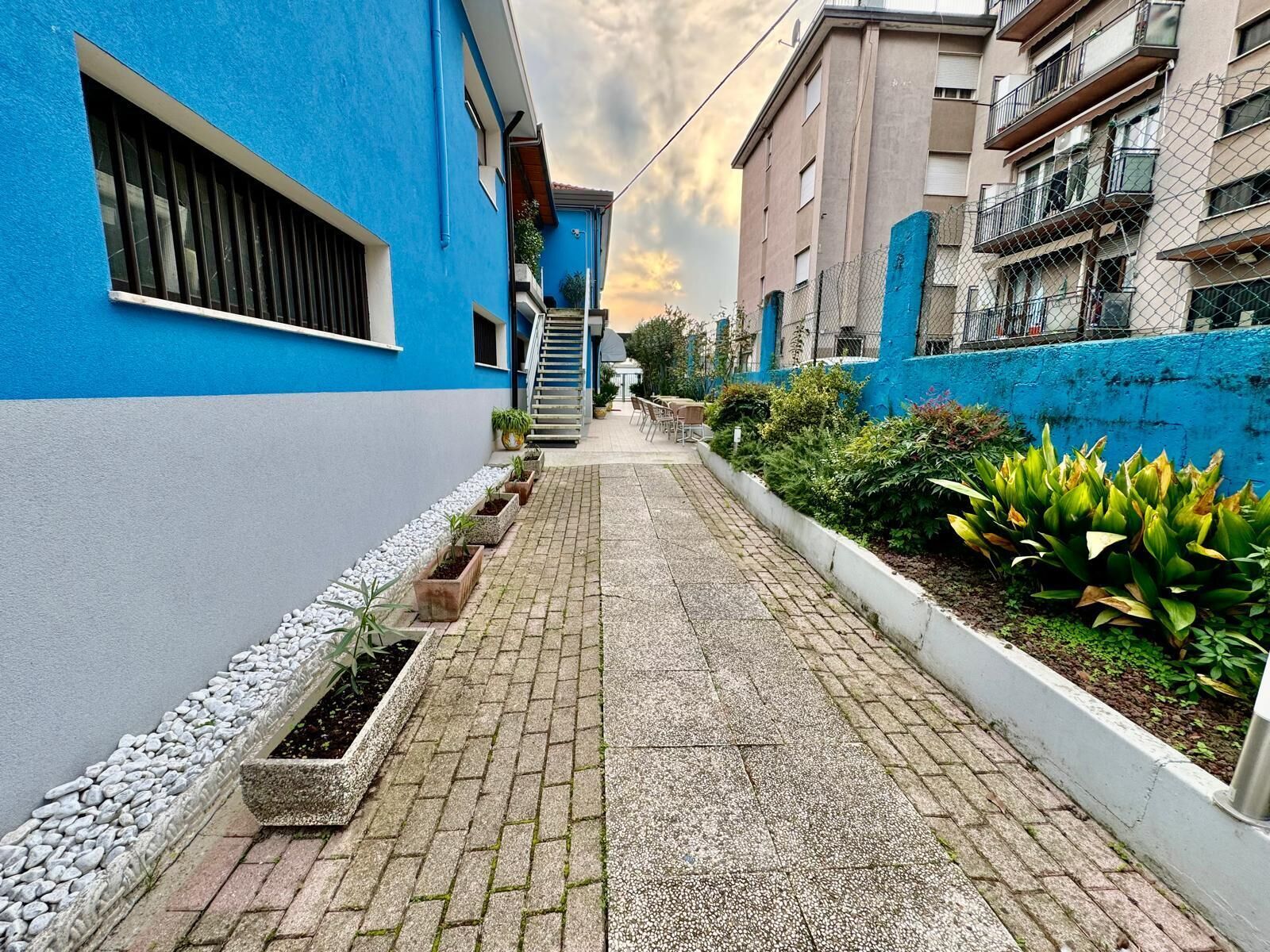 A narrow alleyway with a blue building on the left and a beige building on the right, both with balconies. There are potted plants along the pathway and a small staircase leading to a door.