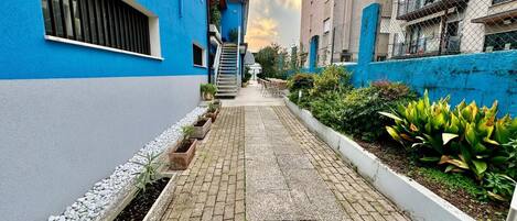 A narrow alleyway with a blue building on the left and a beige building on the right, both with balconies. There are potted plants along the pathway and a small staircase leading to a door.