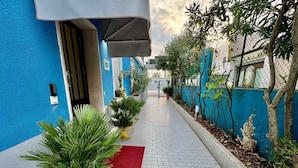 A pathway lined with potted plants leading to a building entrance with a blue wall and a red mat. - Hotel Guidi (Mestre)